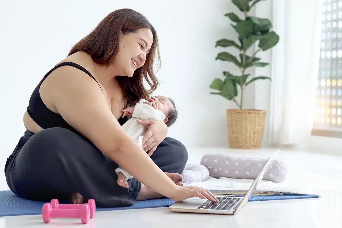 Plus-size mother holding baby, using laptop.